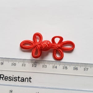 K&ouml;nnte beinhalten: Ein leuchtend roter chinesischer Knebelknopf aus geflochtener Kordel mit einem zentralen Knoten und zwei symmetrischen Schlaufen auf jeder Seite. Ein transparentes Lineal liegt darunter und zeigt, dass der Verschluss etwa 3,8 cm misst.