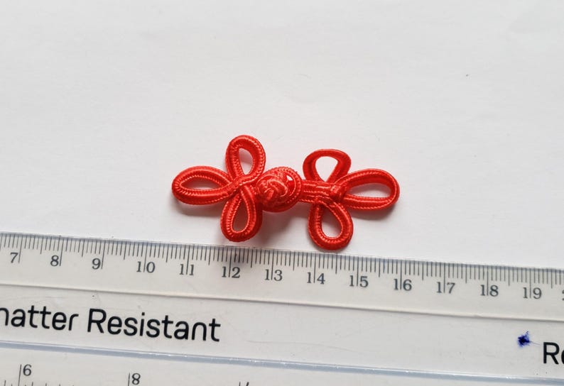 K&ouml;nnte beinhalten: Ein leuchtend roter chinesischer Knebelknopf aus geflochtener Kordel mit einem zentralen Knoten und zwei symmetrischen Schlaufen auf jeder Seite. Ein transparentes Lineal liegt darunter und zeigt, dass der Verschluss etwa 3,8 cm misst.