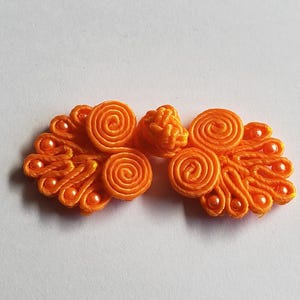 K&ouml;nnte beinhalten: Ein leuchtend orangefarbener Knebelverschluss mit komplizierten, kordelartigen Spiralmustern und kleinen passenden Perlen. Der Verschluss besteht aus zwei symmetrischen Seiten mit gewickelten Stoffschlaufen und einem zentralen Knoten.