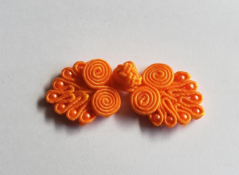 K&ouml;nnte beinhalten: Ein leuchtend orangefarbener Knebelverschluss mit komplizierten, kordelartigen Spiralmustern und kleinen passenden Perlen. Der Verschluss besteht aus zwei symmetrischen Seiten mit gewickelten Stoffschlaufen und einem zentralen Knoten.