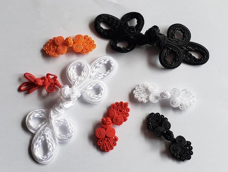 K&ouml;nnte beinhalten: Eine Sammlung dekorativer chinesischer Knebelverschl&uuml;sse in verschiedenen Farben und Gr&ouml;&szlig;en. Das Set enth&auml;lt orange, schwarze, wei&szlig;e und rote geknotete Kordelverschl&uuml;sse mit komplexen Schlaufenmustern, die verstreut auf einer Fl&auml;che angeordnet sind.