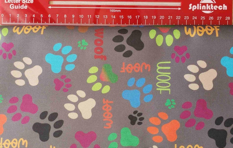 May include: Gray fabric with a repeating pattern of colorful paw prints and the word "woof" in various colors. The paw prints are in shades of pink, black, white, blue, green, and orange. A red ruler is at the top.