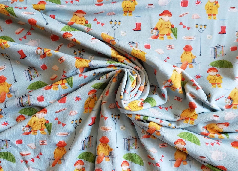 May include: Light blue fabric with a repeating pattern of a bear wearing a yellow raincoat and red hat, holding a lantern and umbrella. The design includes red boots, flags, and teacups.