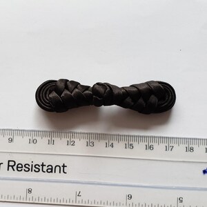 K&ouml;nnte beinhalten: Ein schwarzer Knebelverschluss aus Stoff mit geflochtenem Design und abgerundeten Enden. Der Verschluss liegt &uuml;ber einem durchsichtigen Kunststofflineal, das eine L&auml;nge von etwa 6,5 cm f&uuml;r dieses N&auml;hzubeh&ouml;rteil anzeigt.