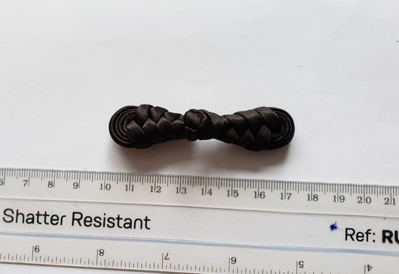 K&ouml;nnte beinhalten: Ein schwarzer Knebelverschluss aus Stoff mit geflochtenem Design und abgerundeten Enden. Der Verschluss liegt &uuml;ber einem durchsichtigen Kunststofflineal, das eine L&auml;nge von etwa 6,5 cm f&uuml;r dieses N&auml;hzubeh&ouml;rteil anzeigt.