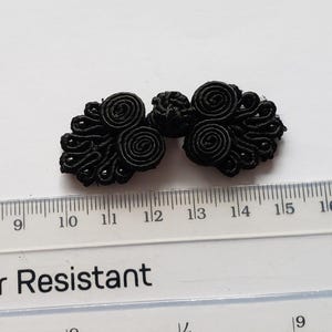 K&ouml;nnte beinhalten: Ein schwarzer Knebelverschluss aus Stoff mit komplexen, spiralf&ouml;rmigen Kordelmustern und einem zentralen Knoten. Der Verschluss liegt &uuml;ber einem durchsichtigen Kunststofflineal und zeigt eine Breite von etwa 3,8 cm von Kante zu Kante.