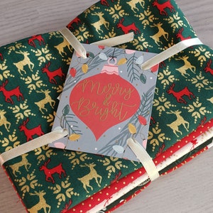 K&ouml;nnte beinhalten: Ein Stapel von vier Stoffquadraten mit gr&uuml;nem Hintergrund und einem Muster aus goldenen und roten Rentieren. Der Stoff ist mit einem wei&szlig;en Band und einem Etikett mit der Aufschrift "Merry & Bright" zusammengebunden.