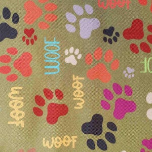 May include: Close-up of a fabric with a repeating pattern of colorful paw prints and the word "Woof" in various colors. The paw prints are in shades of red, orange, purple, and black on a green background.