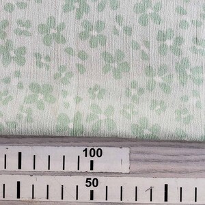 Pu&ograve; includere: Primo piano di un tessuto bianco con un motivo floreale verde. Un righello &egrave; visibile nella parte inferiore dell'immagine con segni per 50, 60 e 100.