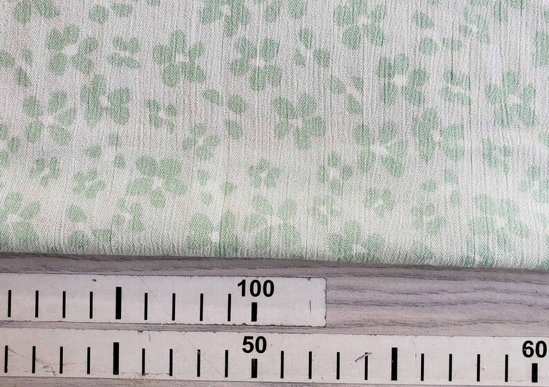 Pu&ograve; includere: Primo piano di un tessuto bianco con un motivo floreale verde. Un righello &egrave; visibile nella parte inferiore dell'immagine con segni per 50, 60 e 100.