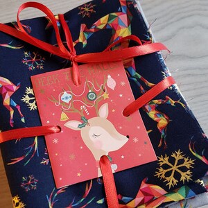 K&ouml;nnte beinhalten: Ein Stapel Stoffquadrate mit einem Rentiermuster in Blaut&ouml;nen, Gold und Regenbogenfarben. Der Stoff ist mit einem roten Band und einer Weihnachtskarte mit einer Rentierillustration und dem Text "Deck the Halls" gebunden.