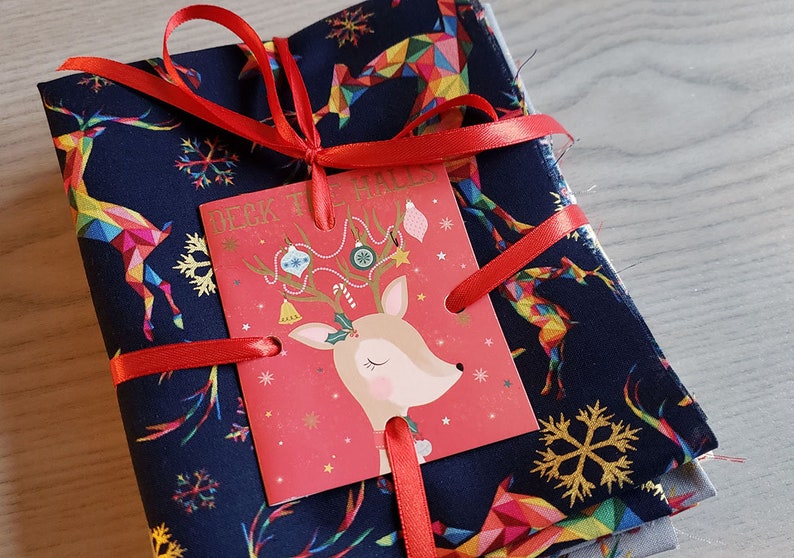 K&ouml;nnte beinhalten: Ein Stapel Stoffquadrate mit einem Rentiermuster in Blaut&ouml;nen, Gold und Regenbogenfarben. Der Stoff ist mit einem roten Band und einer Weihnachtskarte mit einer Rentierillustration und dem Text "Deck the Halls" gebunden.
