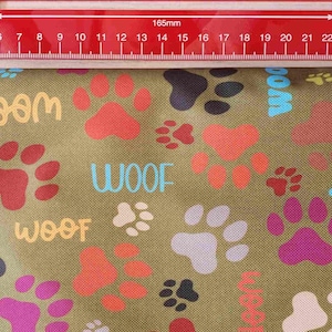 May include: Olive green fabric with a repeating pattern of colorful paw prints and the word "WOOF" in various colors. A red ruler is placed across the top of the fabric, measuring 165mm.