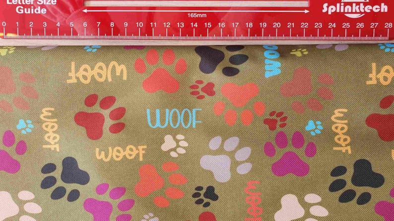 May include: Olive green fabric with a repeating pattern of colorful paw prints and the word "WOOF" in various colors. A red ruler is placed across the top of the fabric, measuring 165mm.