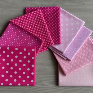 May include: A collection of folded fabric squares arranged in a fan shape, featuring various shades of pink ranging from deep magenta to light pastel. Each piece of fabric displays a white polka dot pattern of varying sizes across the surface.
