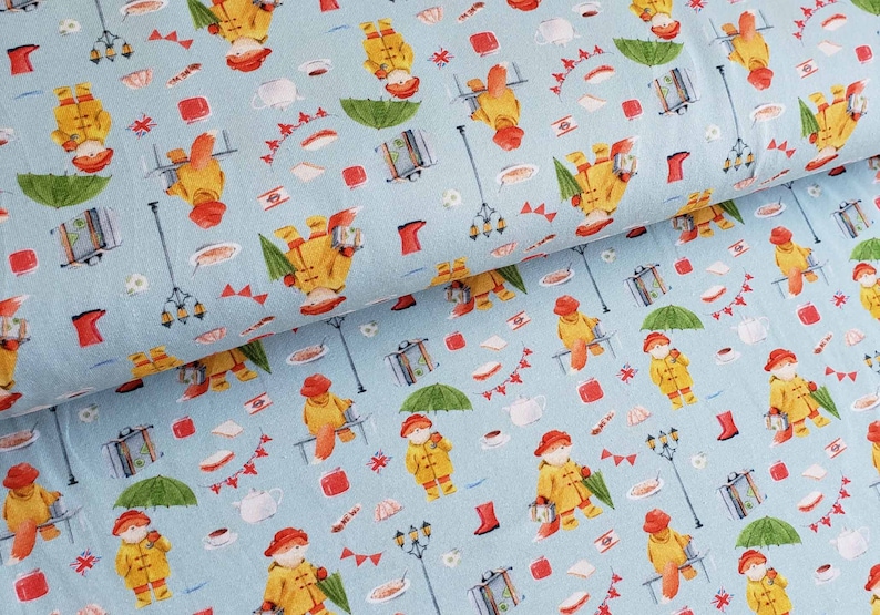 May include: Light blue fabric with a repeating pattern of a bear in a yellow raincoat, red hat, and boots. The design includes umbrellas, suitcases, teacups, and street lamps. The fabric is suitable for various sewing projects.