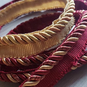 May include: Close-up of decorative trim in shades of burgundy and gold. The trim features a twisted rope design along the edge, with a woven fabric base. The colors and textures suggest a luxurious or vintage aesthetic, suitable for upholstery or craft projects.