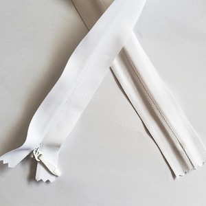 May include: A white zipper with a silver pull tab, lying diagonally across a white surface. The zipper teeth are visible, and the fabric tape is a matching white color. The zipper is open, showing the separation of the two sides.