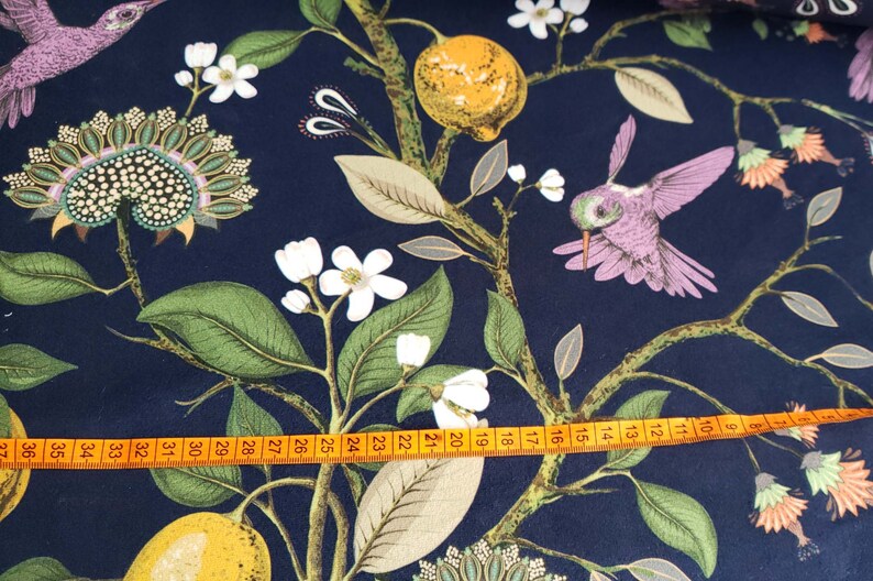 May include: A navy blue fabric with a repeating pattern of hummingbirds, lemons, and white flowers. A measuring tape is placed across the bottom of the fabric.