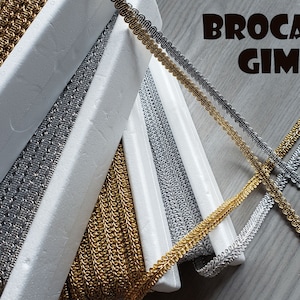 May include: Four spools of gold and silver brocade trim, also known as gimp, are arranged in a criss-cross pattern on a woodgrain surface. The trim is a decorative, woven fabric used for embellishing garments and home decor.