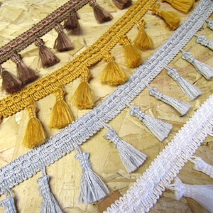 May include: Four different trims with tassels in brown, gold, silver, and white. The trims are made of woven fabric and have a fringe of tassels.
