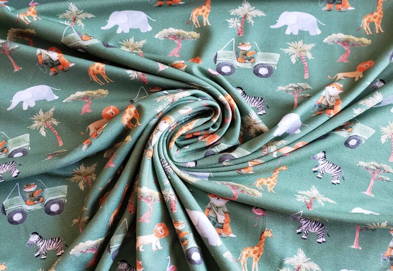 May include: Green fabric with a repeating pattern of safari animals, including elephants, zebras, giraffes, lions, and a jeep. The fabric is folded and draped, showing the pattern.
