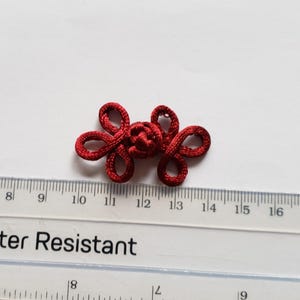 K&ouml;nnte beinhalten: Ein dunkelroter Froschverschluss aus Stoff mit einem zentralen Knoten und vier symmetrischen Schlaufen. Der Verschluss liegt &uuml;ber einem durchsichtigen Kunststofflineal und zeigt eine Breite von etwa 3,8 cm an den Au&szlig;enkanten.