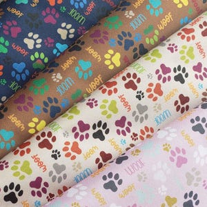 Peut inclure: Gros plan sur des échantillons de tissu avec un motif répété de pattes colorées et le mot "Woof". Les tissus sont dans des tons de bleu, marron, crème et rose, avec le texte "100m" également visible.
