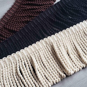 Puede incluir: Tres tiras de flecos decorativos dispuestos en diagonal. Cada tira presenta un encabezado tejido con borlas de cuerda trenzada colgantes. Los colores incluyen un marrón oscuro, un negro sólido y un tono crema blanquecino en primer plano.