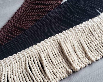 Può includere: Tre strisce di frange decorative disposte in diagonale. Ogni striscia presenta un'intestazione intrecciata con nappe di corda ritorta pendenti. I colori includono un marrone scuro, un nero solido e una tonalità crema bianco sporco in primo piano.