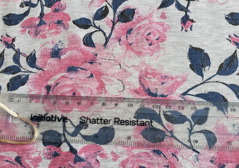 May include: A close-up of a gray fabric with a pink and blue floral print. The fabric has a ruler on top of it with the words "initiative Shatter Resistant" printed on it.