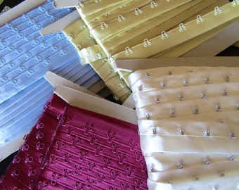 May include: Four spools of bra strap hook and eye tape in different colours: light blue, ivory, burgundy, and beige. The tape is made of satin and has metal hooks and eyes.