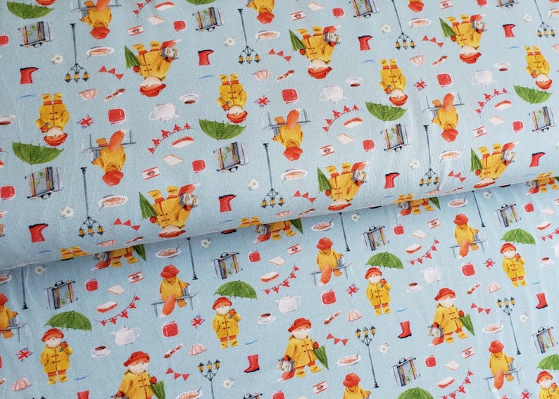 May include: Light blue fabric with a repeating pattern of a bear in a yellow raincoat, red hat, and boots. The design includes umbrellas, suitcases, and street lamps, creating a whimsical, storybook theme.
