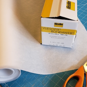 May include: A roll of white fusible web, a box of Vlieseline Bondaweave fusible web, and a pair of orange-handled scissors on a blue cutting mat.