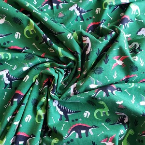 May include: Green fabric with a repeating pattern of black, red, and green dinosaurs. The fabric has a white background with a green pattern.