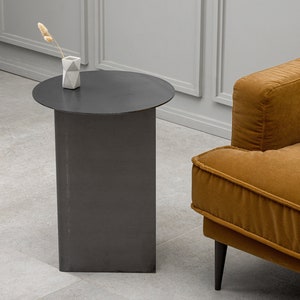 Op de afbeelding: Een ronde zwarte bijzettafel met een cilindrische basis en een kleine witte vaas met een gedroogde bloemstengel erop. De tafel staat naast een bruine fluwelen bank met zwarte poten.