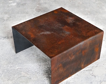 Op de afbeelding: Een vierkante, roestige metalen tafel met een zwarte metalen basis. De tafel is gemaakt van één stuk metaal dat in vorm is gebogen.