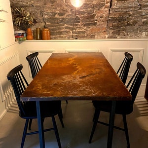 May include: A rustic dining table with a brown, textured, metal top and a black metal frame. Four black chairs surround the table. The table is set against a stone wall.