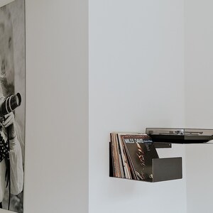 May include: A black wall shelf with a record player and vinyl records. The shelf is mounted on a white wall. A black and white photo of a woman holding a camera is hanging on the wall.