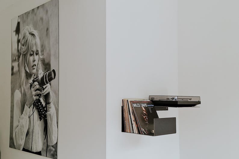 May include: A black wall shelf with a record player and vinyl records. The shelf is mounted on a white wall. A black and white photo of a woman holding a camera is hanging on the wall.
