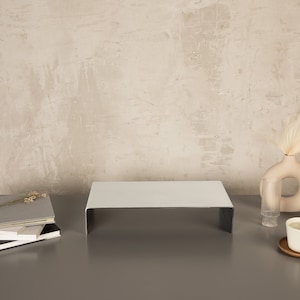 Pu&ograve; includere: Un supporto in metallo bianco con una superficie superiore piatta, una pila di libri, un bicchiere trasparente, un vaso beige con fiori secchi e una tazza in ceramica bianca su un sottobicchiere di legno su una superficie grigia.
