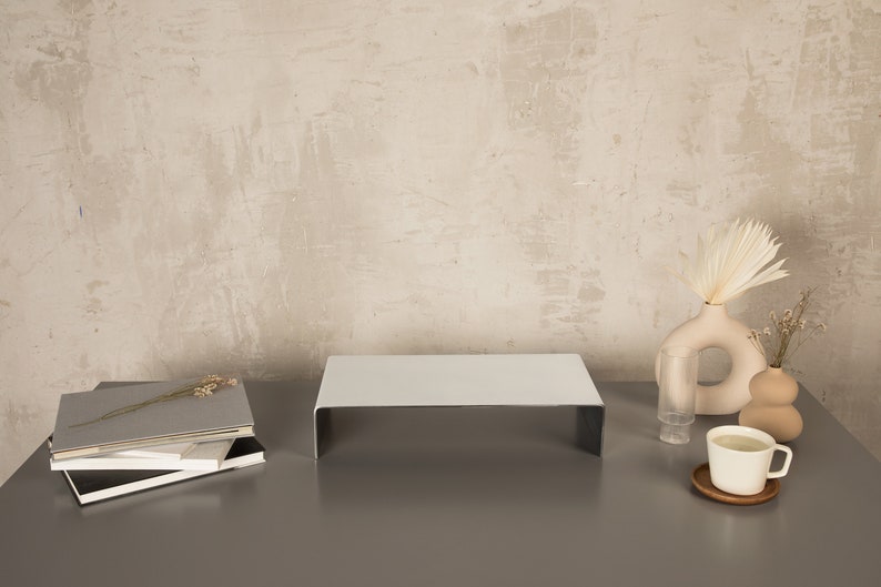 Pu&ograve; includere: Un supporto in metallo bianco con una superficie superiore piatta, una pila di libri, un bicchiere trasparente, un vaso beige con fiori secchi e una tazza in ceramica bianca su un sottobicchiere di legno su una superficie grigia.