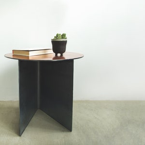 Op de afbeelding: Een kleine, ronde, koperkleurige bijzettafel met een zwarte metalen basis. Een boek en een plant in een pot staan op de tafel.
