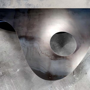 May include: A large, abstract metal sculpture with a circular cutout. The sculpture is made of a dark, metallic material and has a smooth, polished finish. The sculpture is resting on a concrete surface.