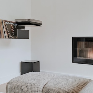 May include: A white room with a fireplace, a record player, and a gray blanket on the floor. The fireplace has a black frame and a glass front with a fire burning inside. The record player is mounted on the wall and has a black shelf with a record on it. The record is by Miles Davis.