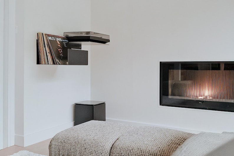 May include: A white room with a fireplace, a record player, and a gray blanket on the floor. The fireplace has a black frame and a glass front with a fire burning inside. The record player is mounted on the wall and has a black shelf with a record on it. The record is by Miles Davis.