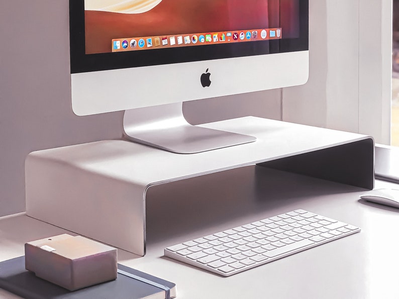 Pu&ograve; includere: Un supporto per monitor in metallo bianco con un bordo anteriore curvo. Una tastiera wireless bianca e una piccola scatola grigia sono sulla scrivania davanti al supporto. Un computer iMac argentato &egrave; posizionato sul supporto.