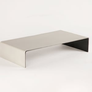 Pu&ograve; includere: Un tavolino da caff&egrave; in metallo grigio chiaro con un piano rettangolare e una base aperta curva. Il tavolo &egrave; realizzato in un unico pezzo di metallo e ha un design moderno e minimalista.
