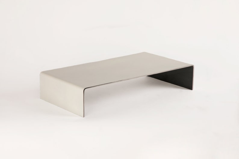 Pu&ograve; includere: Un tavolino da caff&egrave; in metallo grigio chiaro con un piano rettangolare e una base aperta curva. Il tavolo &egrave; realizzato in un unico pezzo di metallo e ha un design moderno e minimalista.