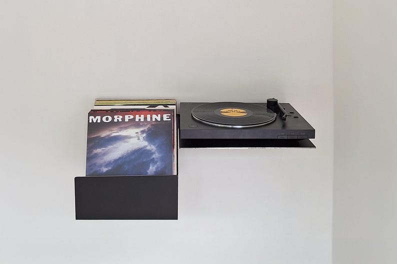 May include: A black record player with a black vinyl record on the turntable. The record player is mounted on a black shelf with a stack of vinyl records underneath. The record cover features the word "MORPHINE" in white text.